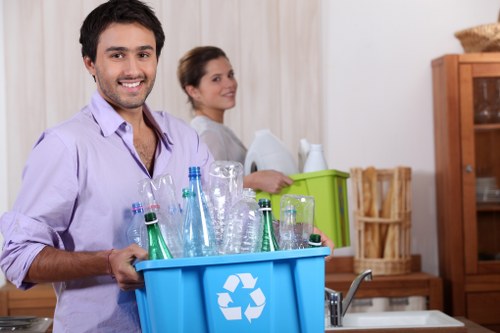 A young man with dark hair and a beard, smiling, is holding a blue recycling bin filled with empty glass bottles and jars, situated in a domestic kitchen environment. Behind him, a woman with dark hair tied back is slightly out of focus, standing near a green plastic crate and a wooden cupboard, both located against a neutral-colored wall. The kitchen features light-colored curtains, a wooden cabinet, and a countertop with a sink, with natural lighting illuminating the scene. The scene suggests a household setting involved in rubbish collection or recycling activities, consistent with the services provided by Garden Clearance Catford in the local area around postcode SE6, including debris and waste removal for domestic properties.