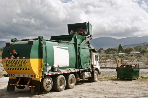 A large green rubbish collection truck with a yellow caution strip and rear warning sign is parked on a gravel or dirt surface at an outdoor disposal site. The truck's rear compactor mechanism is in an open position, suggesting it may be in the process of collecting or dumping waste. Next to the truck, there is a green waste bin filled with what appears to be discarded materials, possibly garden or household waste. The background features a chain-link fence, distant rolling hills, and a cloudy sky indicating overcast weather. The scene is typical of rubbish removal operations and reflects a professional waste collection service, such as Garden Clearance Catford, operating in a rural or semi-rural environment within the UK, with the area possibly near Catford or a similar location in South London.