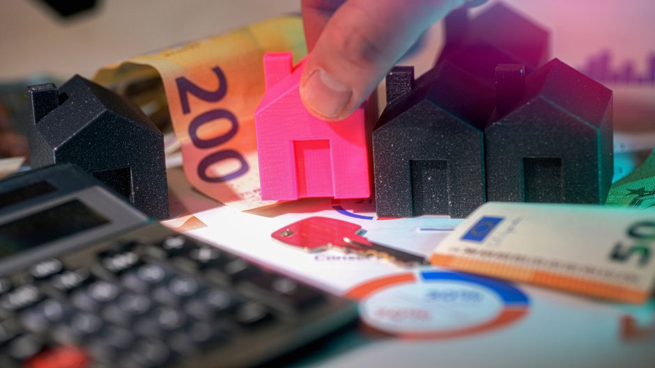 A close-up view of a person’s hand holding a small pink model house positioned among three larger black model houses, all made of textured material resembling foam or plastic, with a backdrop featurin