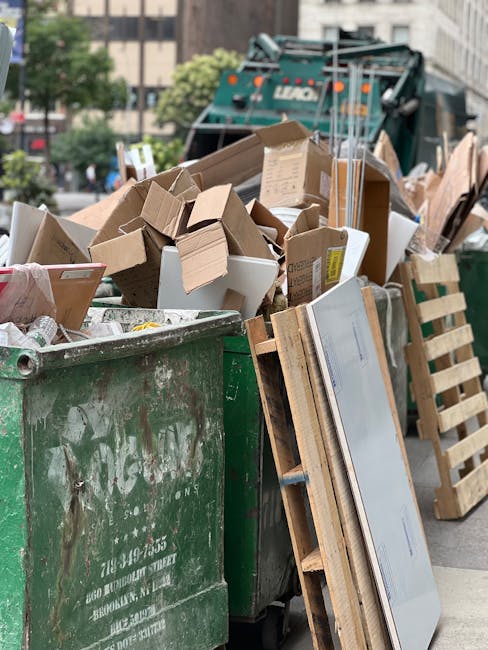 A large green waste skip filled with various types of rubbish, including cardboard boxes, foam packaging, and miscellaneous discarded materials, located outdoors on a paved street. The cardboard boxes