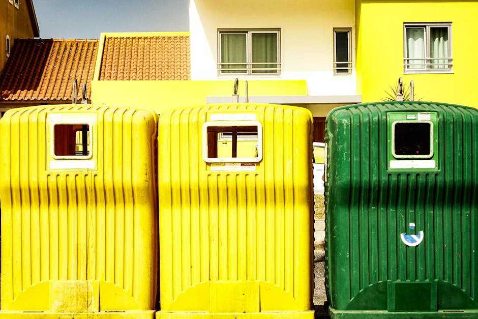 A large green waste skip filled with various types of rubbish, including cardboard boxes, foam packaging, and miscellaneous discarded materials, located outdoors on a paved street. The cardboard boxes